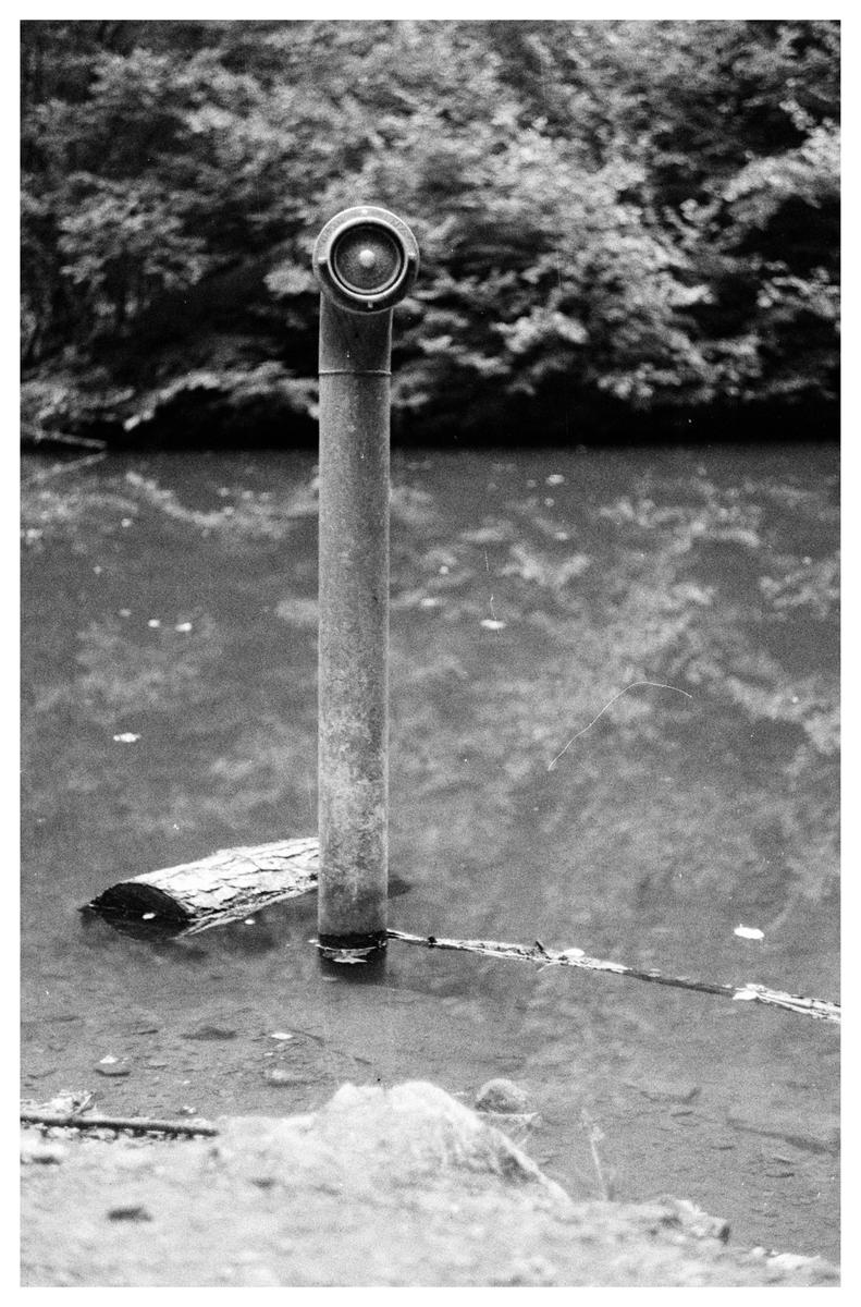 A periscope-like pipe stands in a body of water with trees in the background.