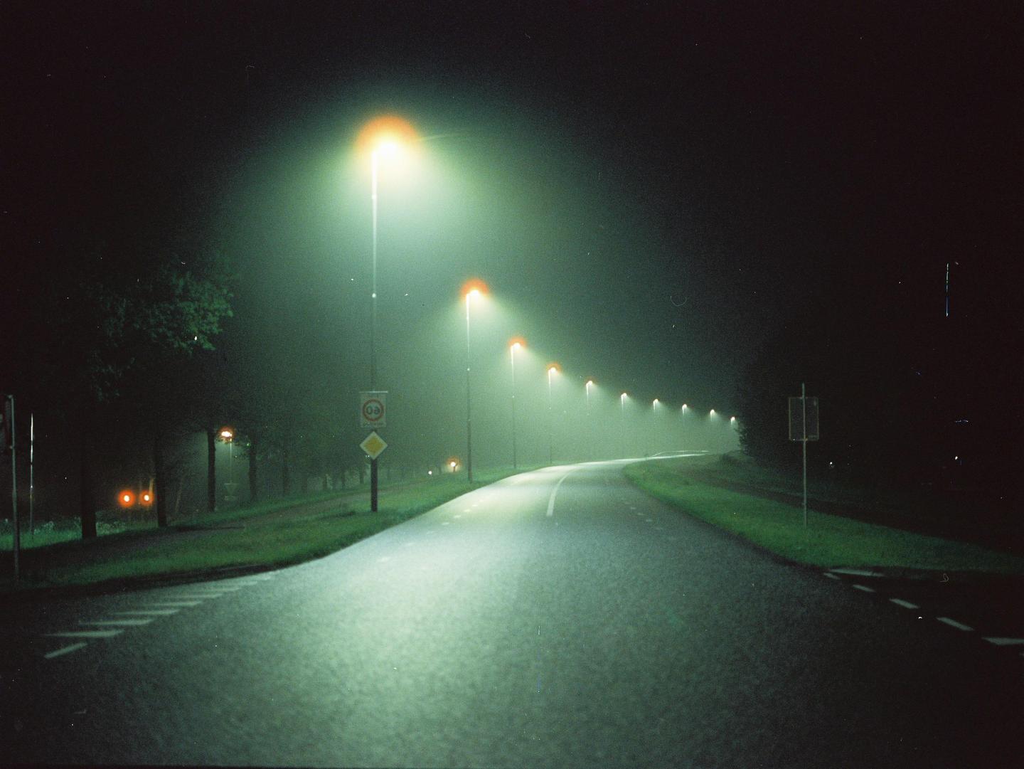 A long, winding road is illuminated by a row of streetlights on a foggy night.