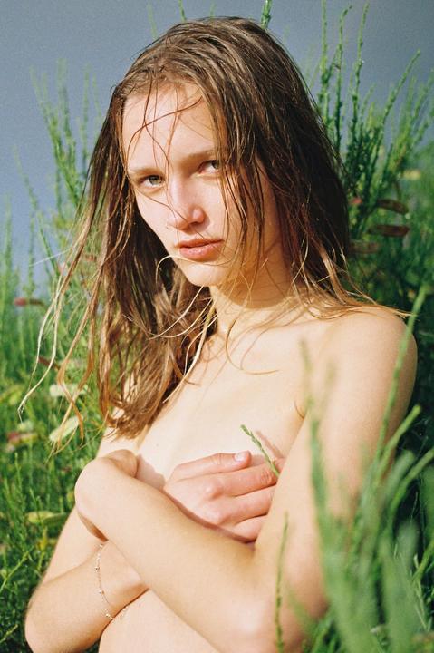 A young woman with wet hair covers her chest with her arms while sitting in tall grass.
