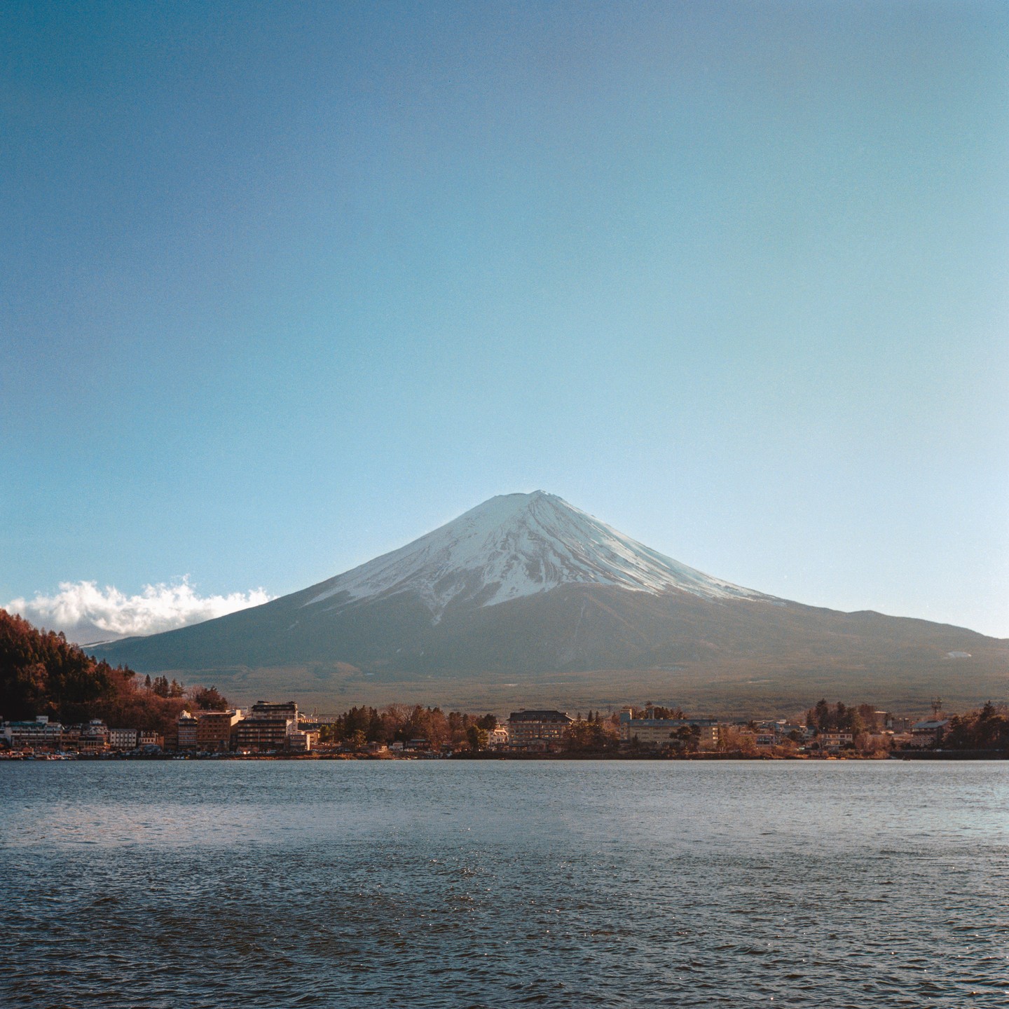 Mount Fuji with a snow-capped peak rises behind a town and a lake.