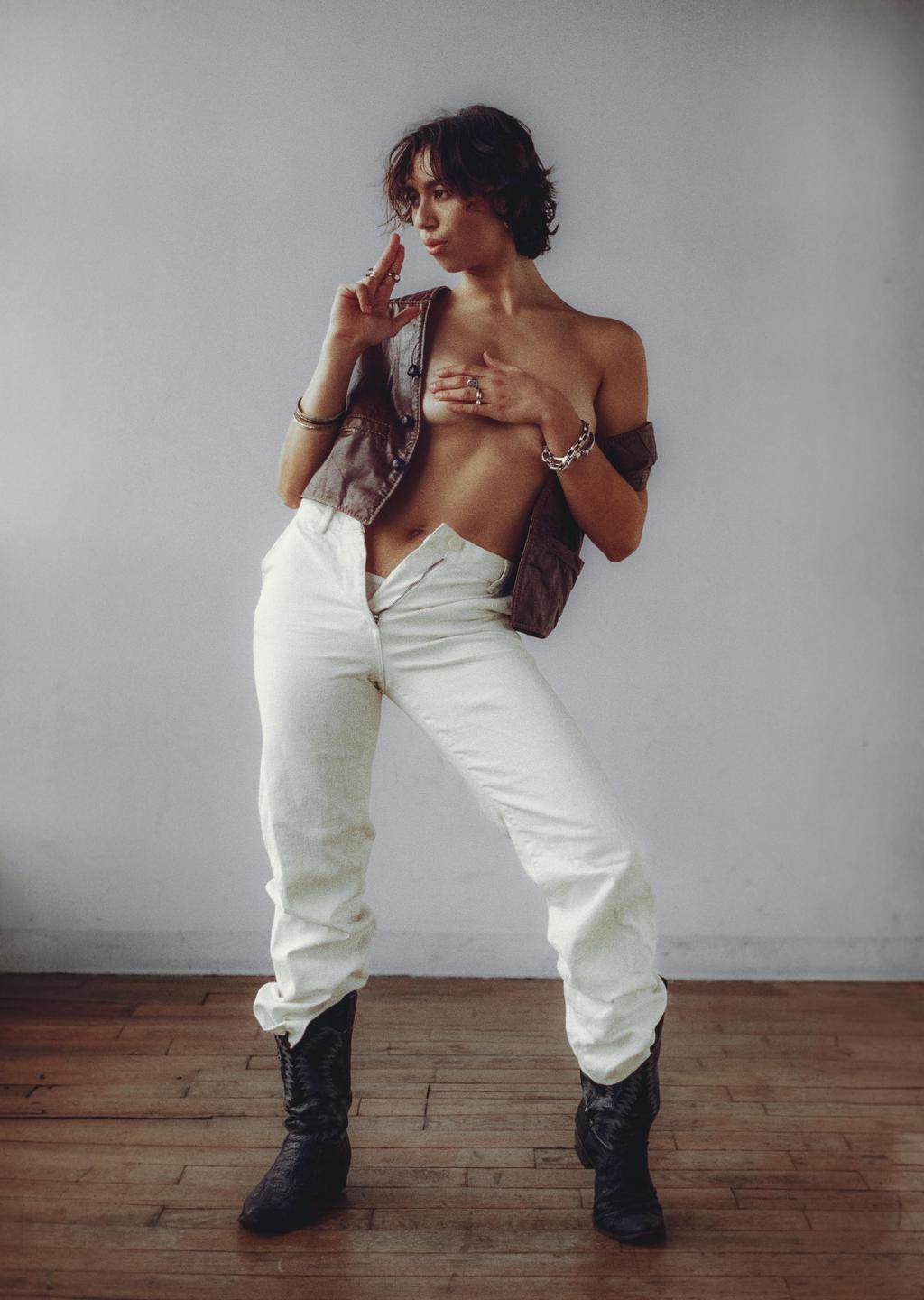 A woman with short hair poses topless in a vest, white pants, and cowboy boots.