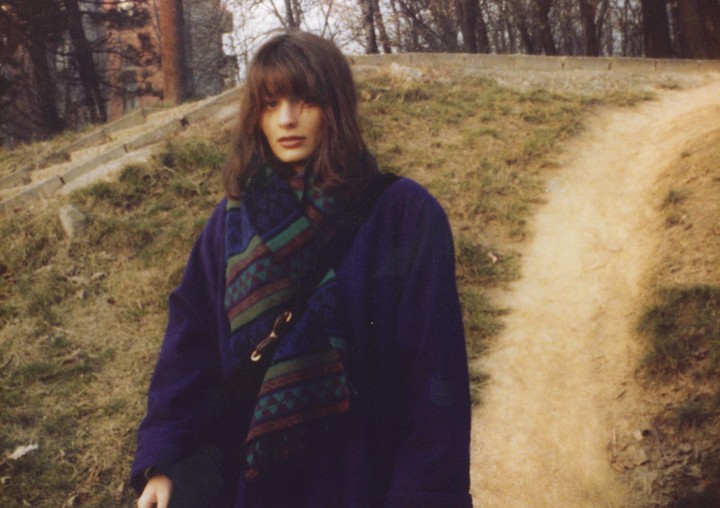 A woman wearing a purple coat and a patterned scarf stands on a grassy hill next to a dirt path.