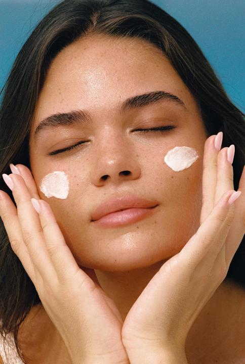 A woman with closed eyes has white cream on her cheeks and hands cupping her face.