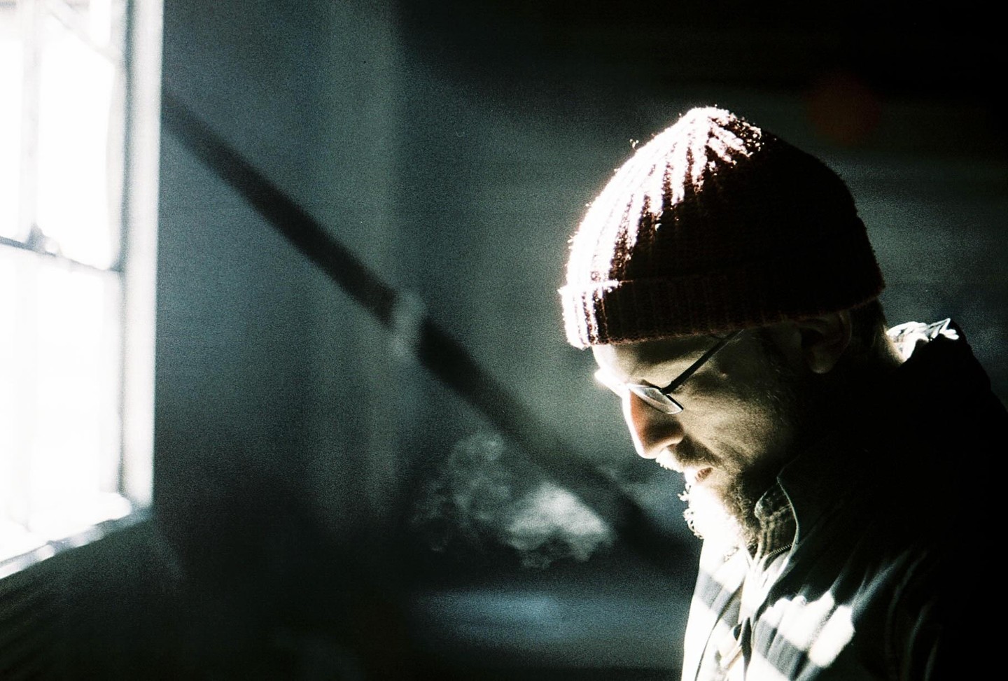 A man in a beanie and glasses is illuminated by a bright light source from the left, with visible breath in the cold air.