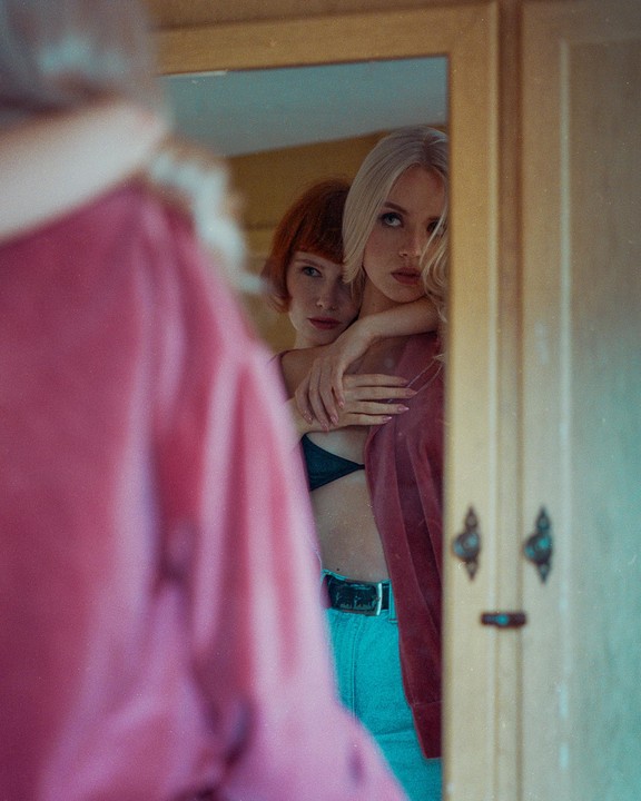Two women are reflected in a mirror, one with red hair and the other with blonde hair, embracing each other.