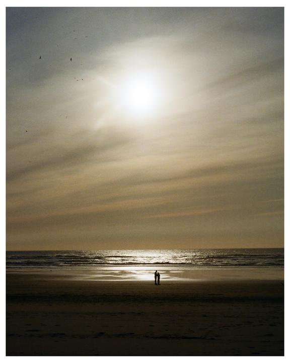 Two people stand on a beach looking out at the ocean under a bright sun.