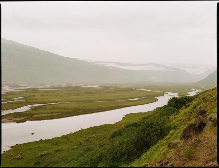 A river winds through a vast green valley with mountains in the distance under a hazy sky.