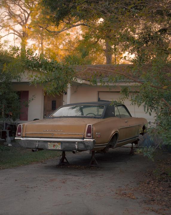 A gold vintage car is parked on stilts in a driveway in front of a house.