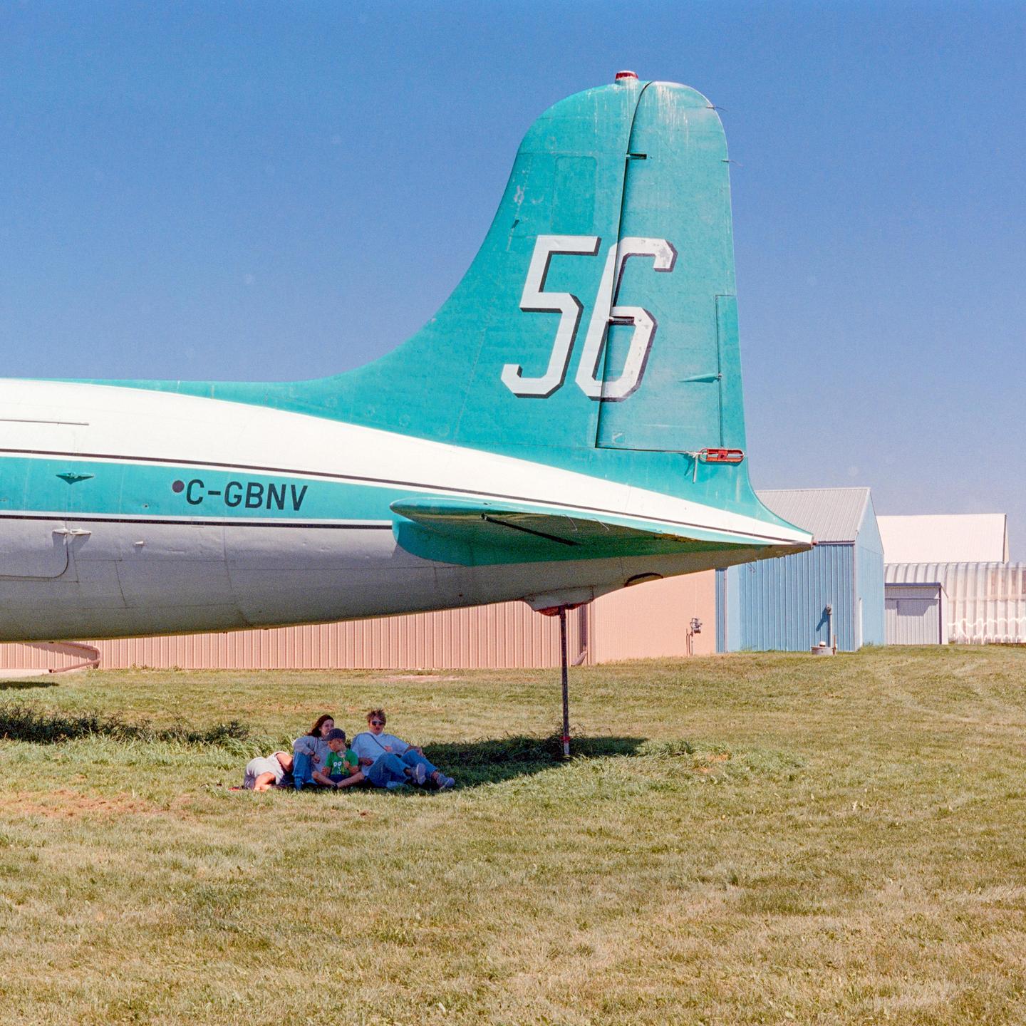 Three people sit in the shade of a large airplane tail on a grassy field.