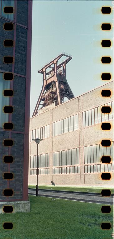 A large industrial structure with a tall metal tower rises above a brick building with many windows, next to a grassy area and a street light.
