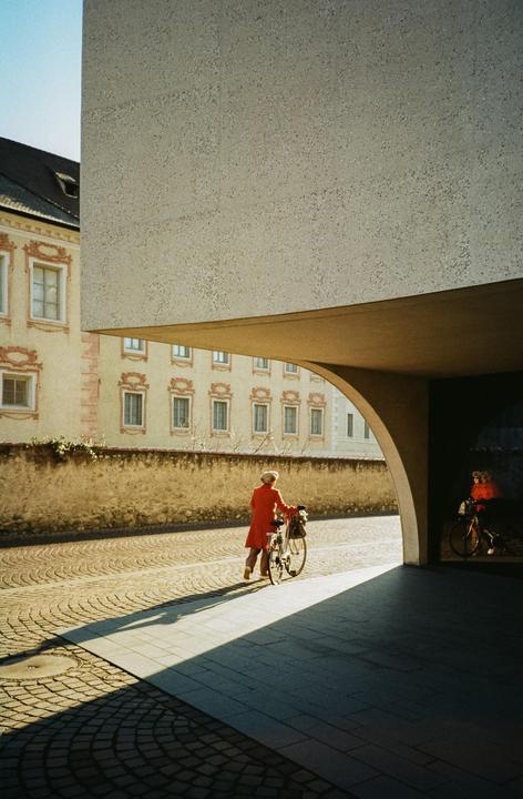 A woman in a red coat walks with her bicycle on a cobblestone street next to a modern building with an arched entrance.