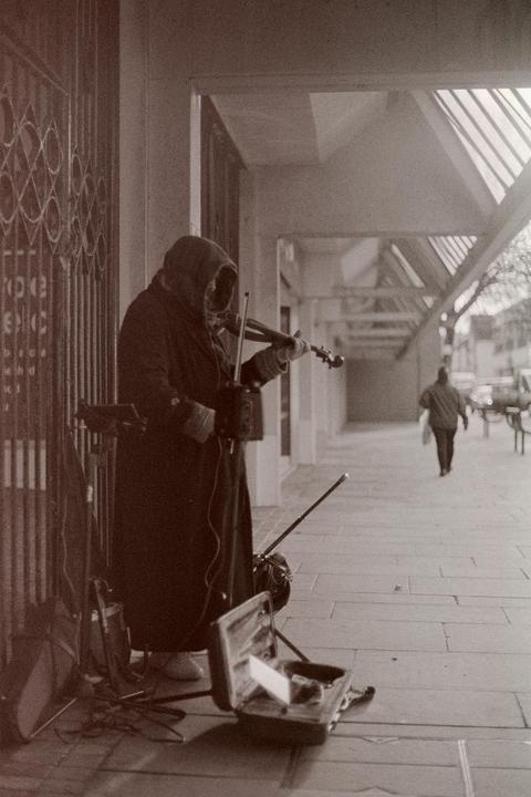 A hooded street musician plays a violin on a sidewalk next to an open violin case.