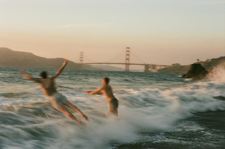 Two men are in the ocean with waves crashing around them, and the Golden Gate Bridge is visible in the background.
