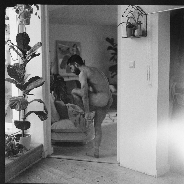 A man is getting dressed in a room with plants and a couch.