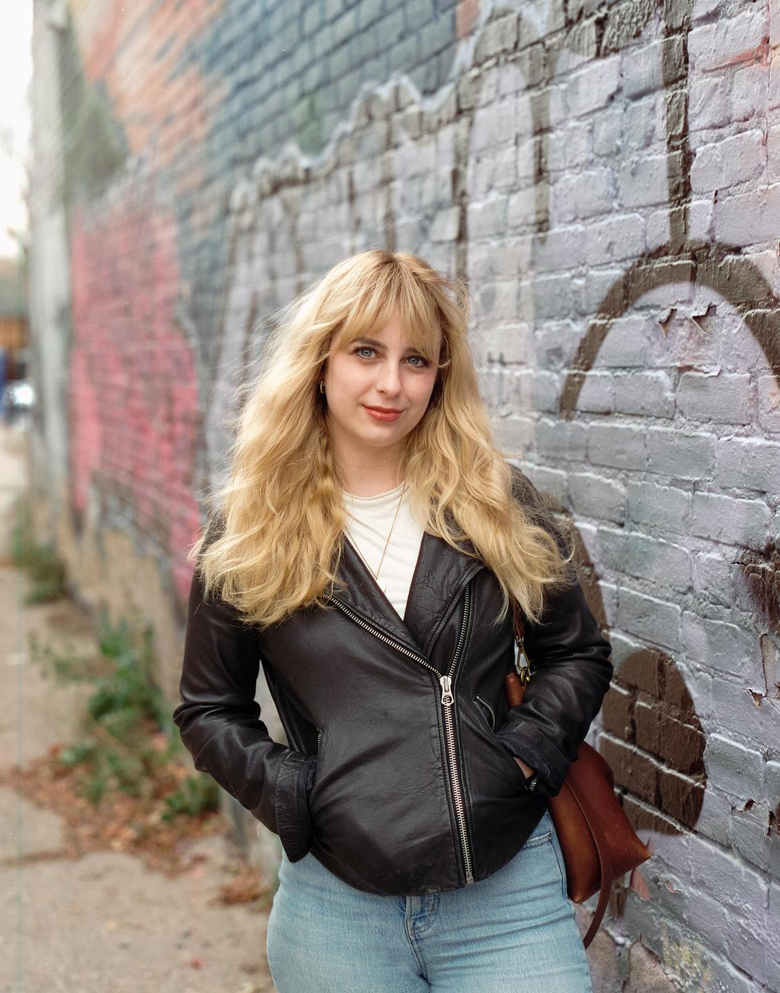 A woman with long blonde hair and a black leather jacket stands in front of a graffiti-covered brick wall.