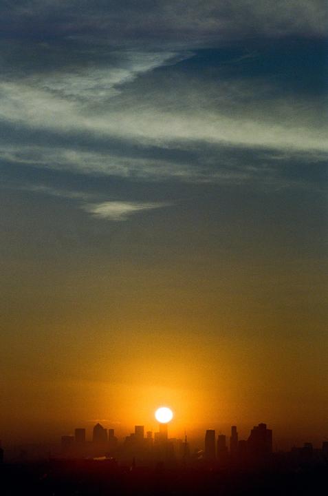The sun rises over the silhouette of a city skyline with clouds in the sky.