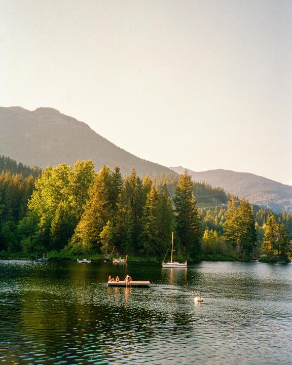 A lake with boats and a floating dock with people is surrounded by a forest and mountains.