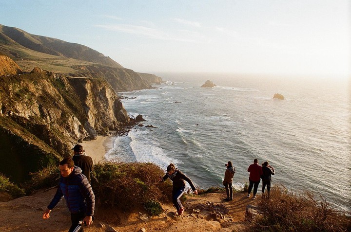 A group of people stand on a cliff overlooking a beach and the ocean with mountains in the background.