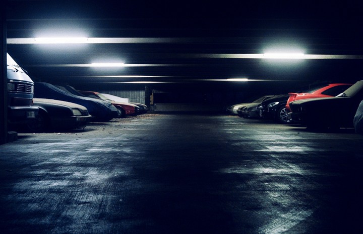 A dark underground parking garage is illuminated by fluorescent lights, revealing rows of parked cars, some covered.