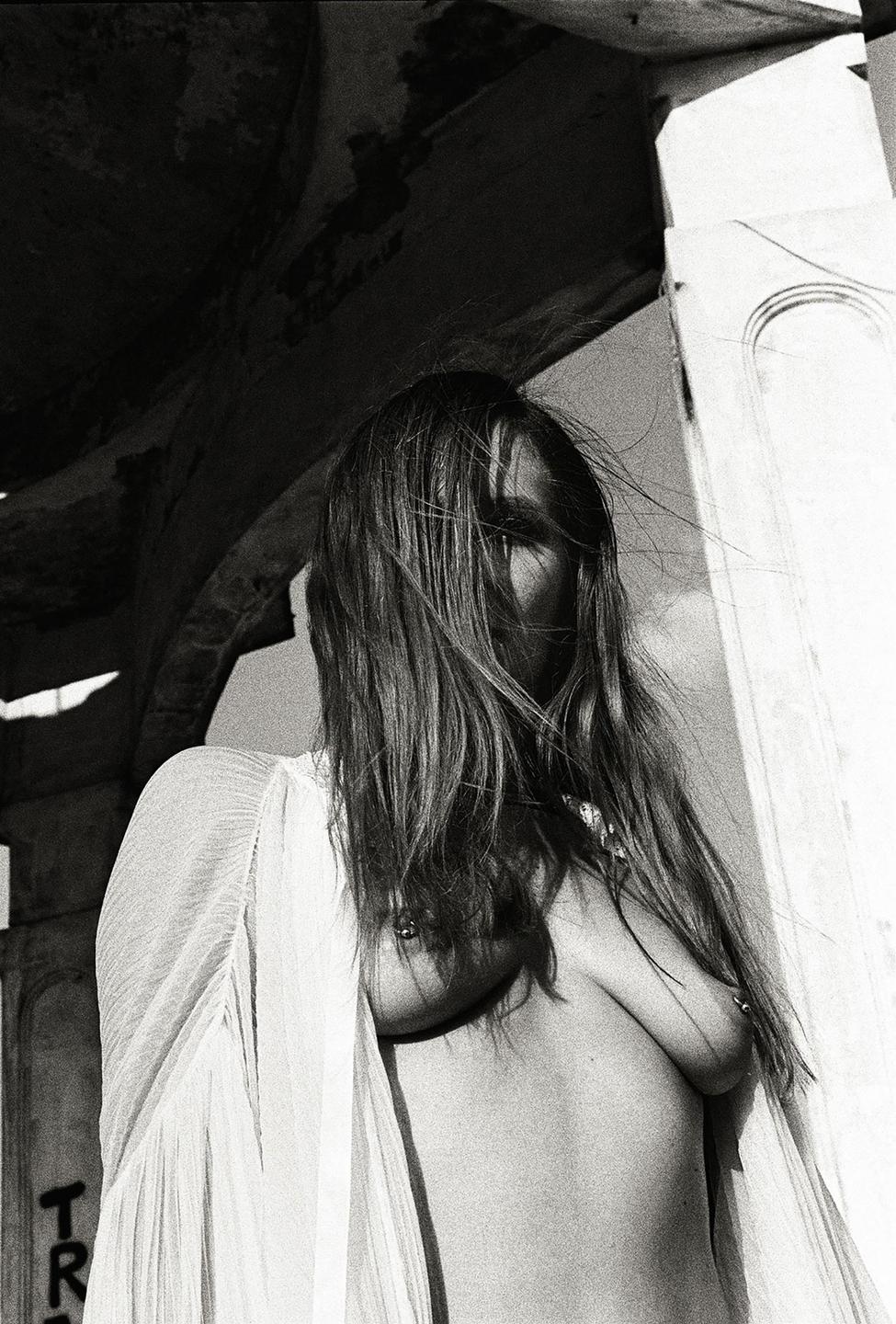 A woman with long hair covering her face stands topless in a sheer top.