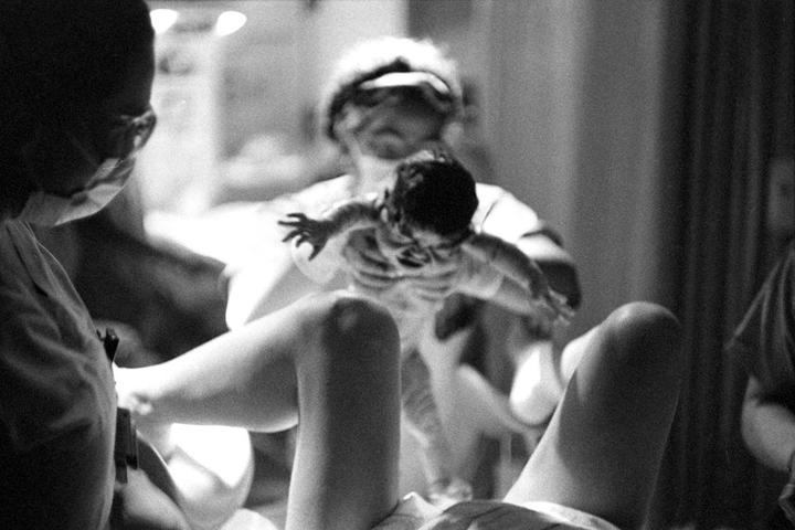 A newborn baby is held up by a medical professional while another medical professional and a woman look on.