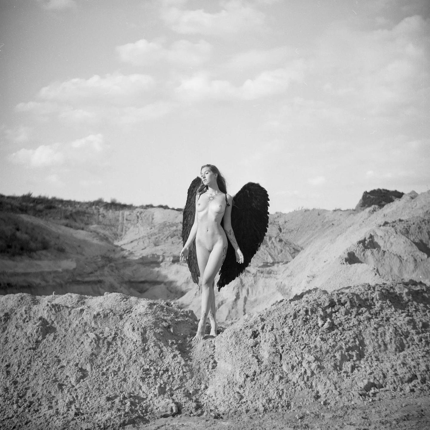 A nude woman with black wings stands on a sandy hill.