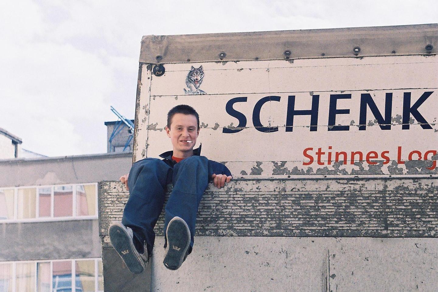 A young man with a cigarette in his mouth sits on the edge of a truck with a wolf sticker on it.