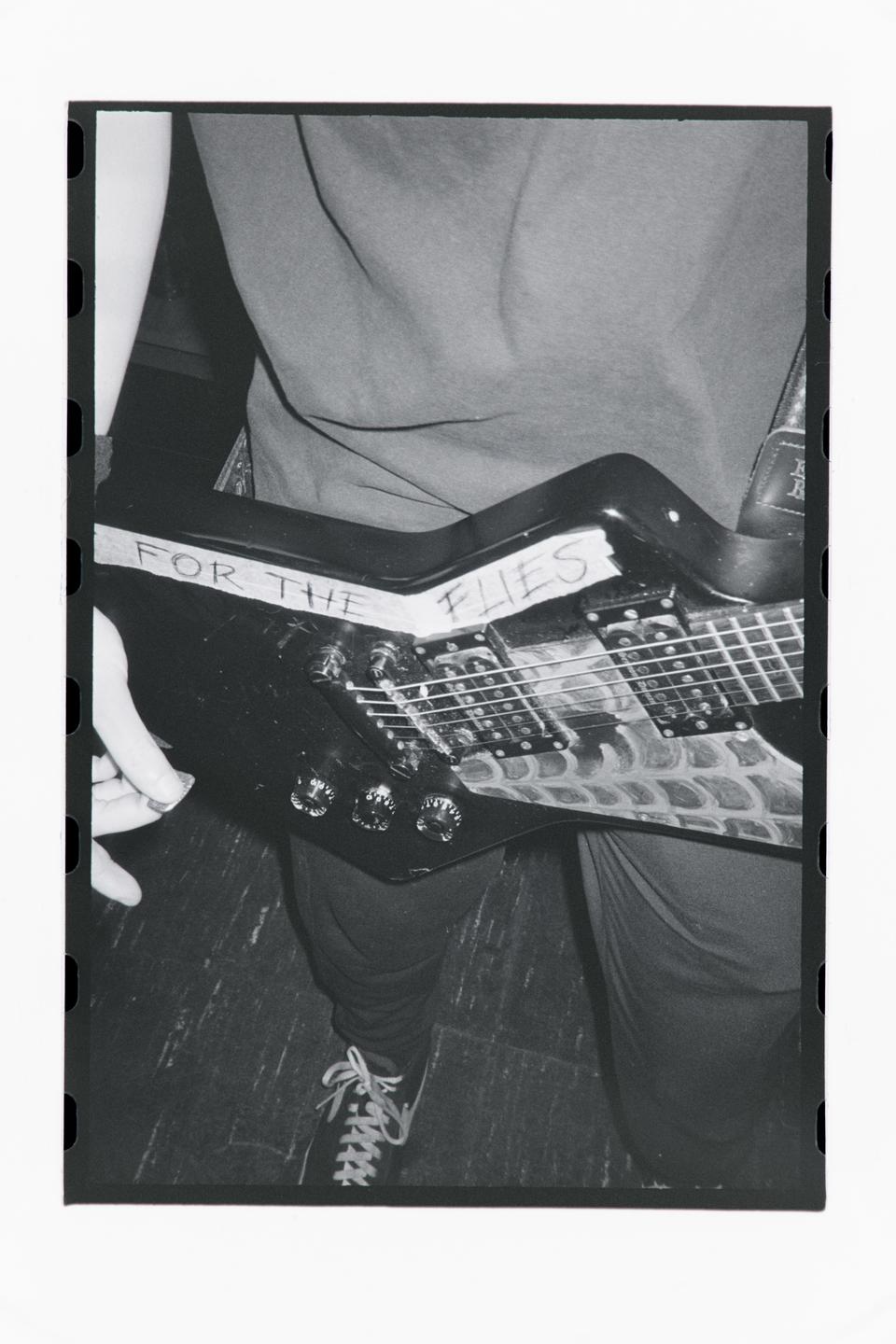 A person holds an electric guitar with "FOR THE FLIES" written on it.