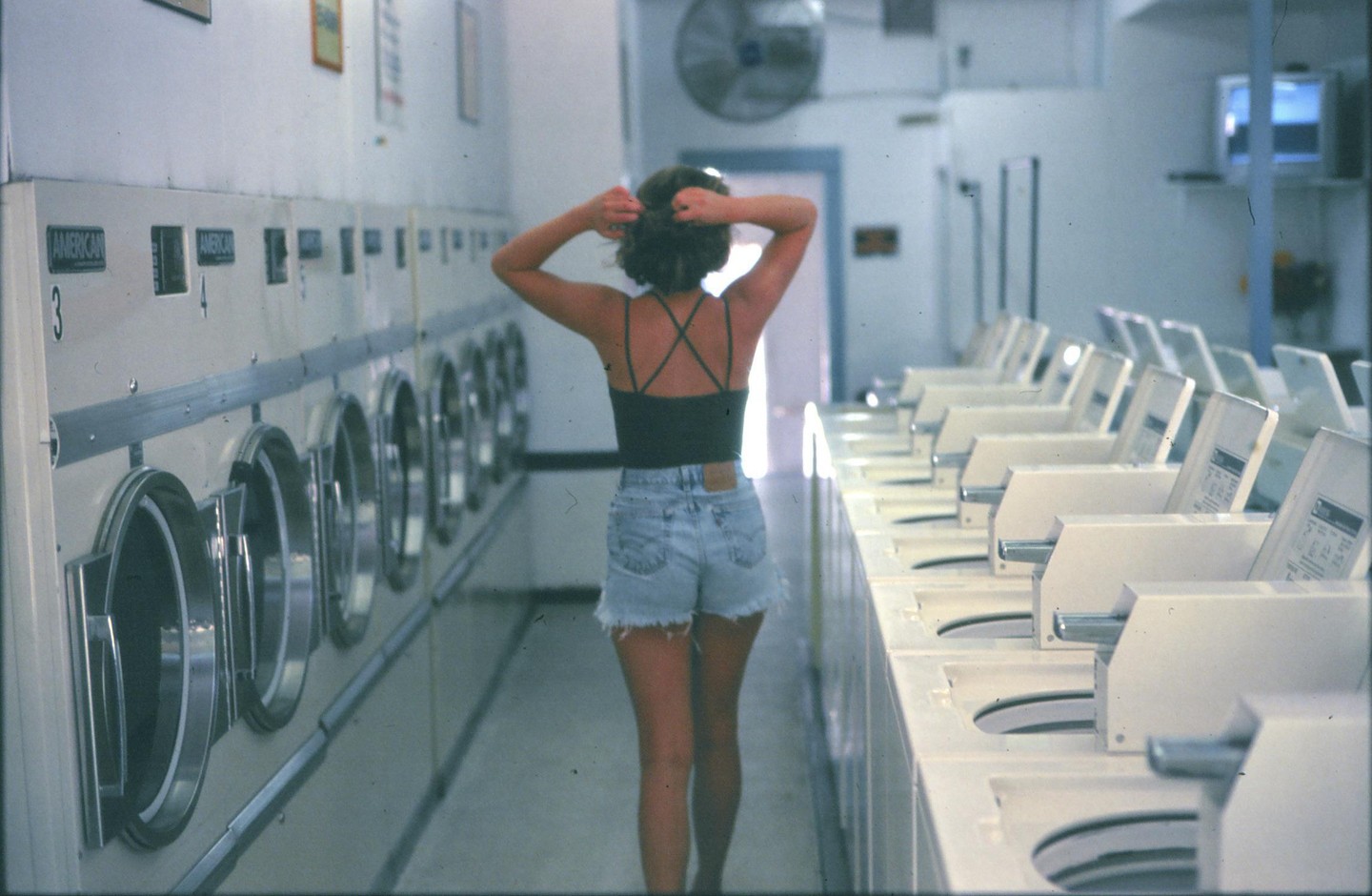A woman stands in a laundromat with washing machines on one side and dryers on the other.