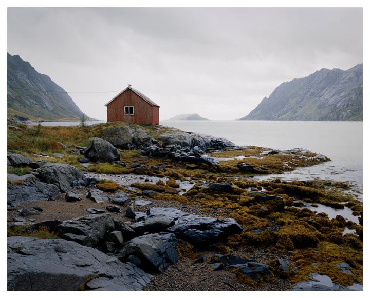 A red cabin sits on a rocky, mossy shore next to a body of water with mountains in the background under a cloudy sky.