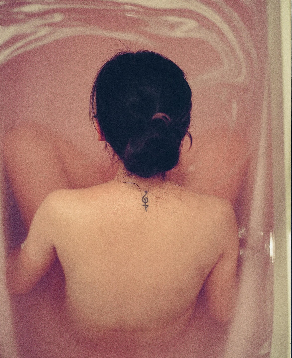 A woman with a bun and a small tattoo on her neck is seen from behind, sitting in a bathtub filled with pink water.