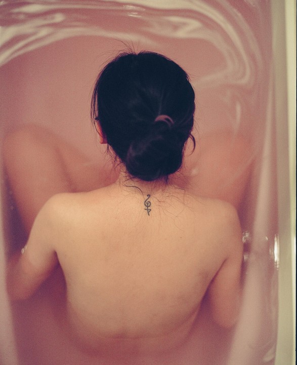 A woman with a bun and a small tattoo on her neck is seen from behind, sitting in a bathtub filled with pink water.