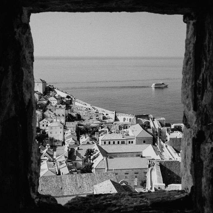 A boat travels on the sea next to a town with many buildings and a wall, all seen through a stone archway.
