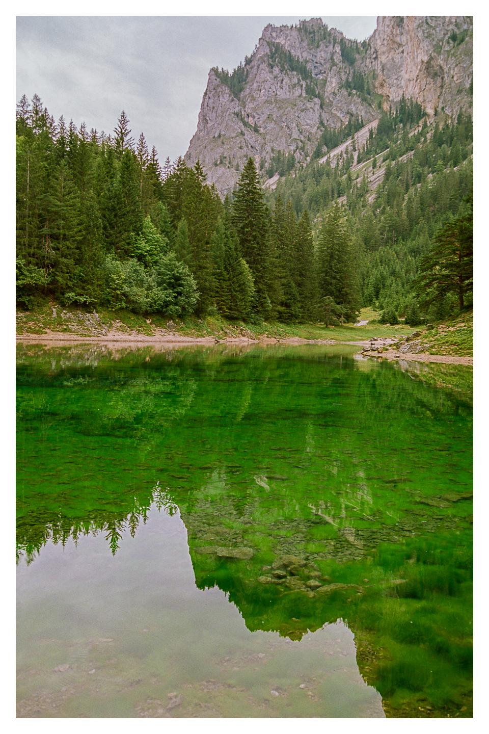 A green lake reflects a mountain and forest under a cloudy sky.