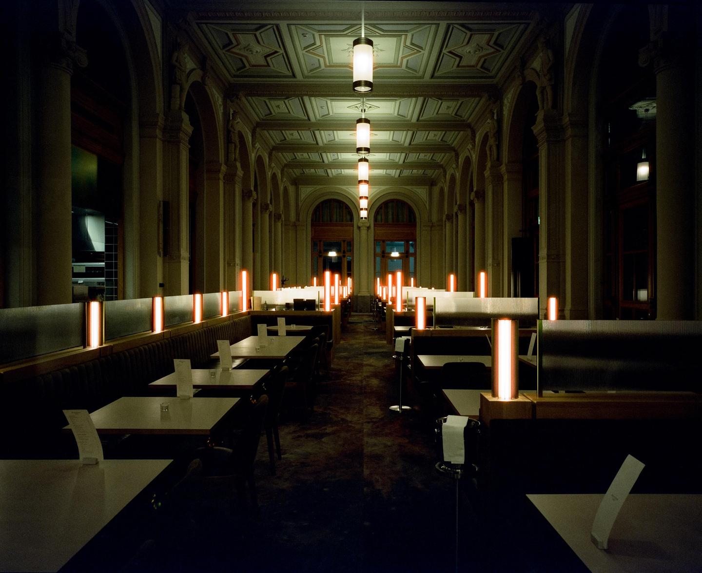 A long, ornate dining hall with many empty tables and booths is illuminated by warm, glowing lights.