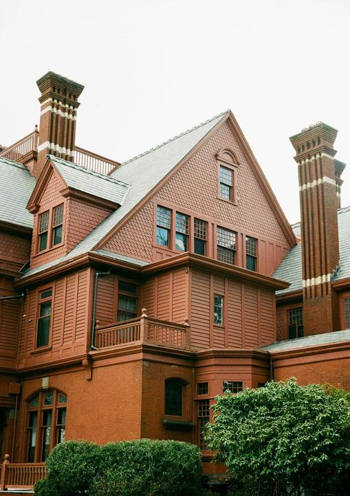 A large, red brick and wood house with multiple gables and chimneys is partially obscured by green bushes.