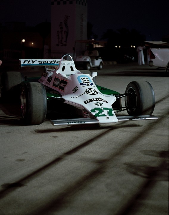 A white and green Formula 1 race car with the number 27 is parked on asphalt at night, casting long shadows.