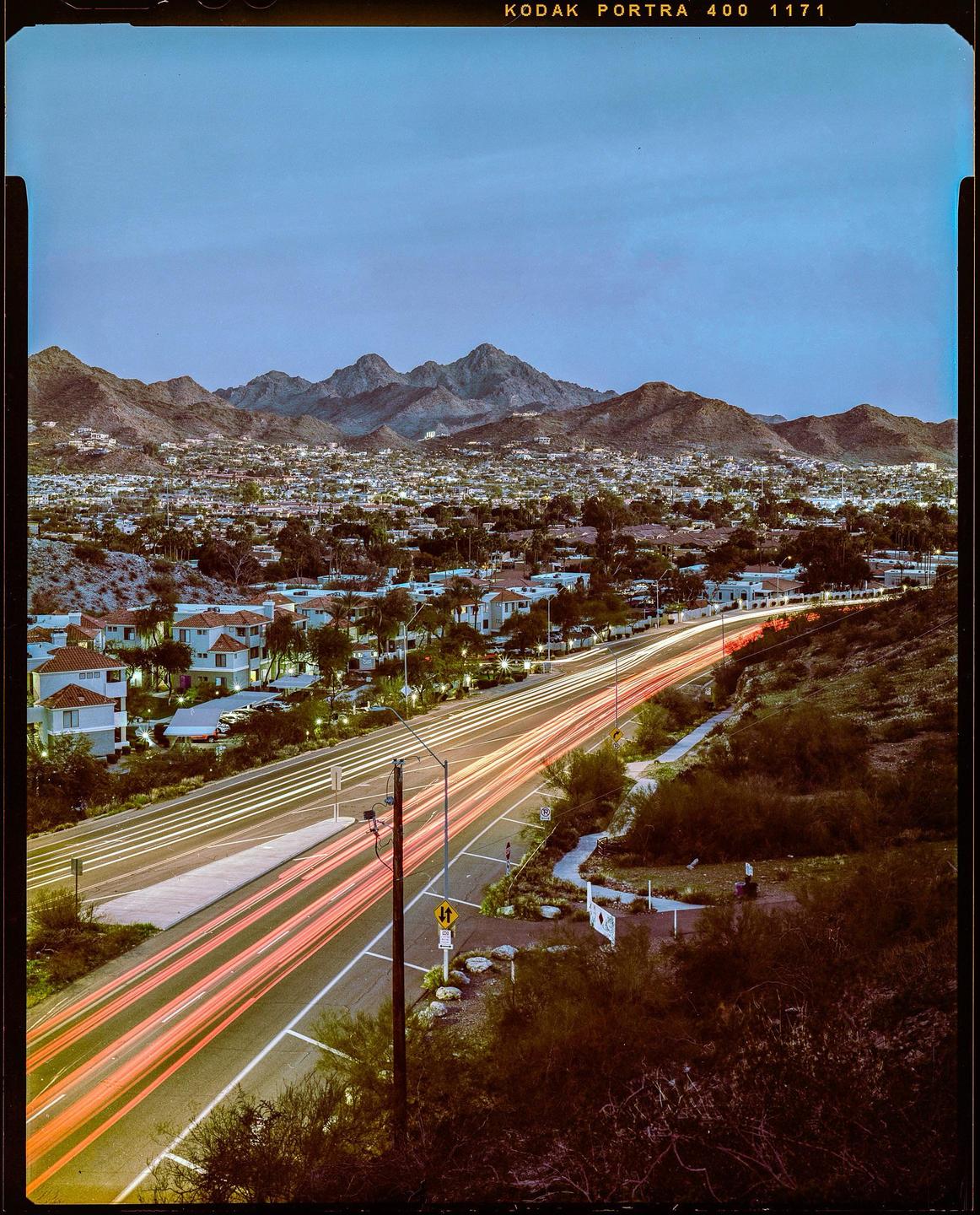 A long exposure captures light trails from cars on a road winding through a town nestled among mountains at dusk.