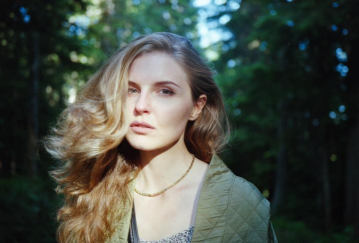 A woman with long, wavy hair and a quilted jacket looks directly forward in a forest.