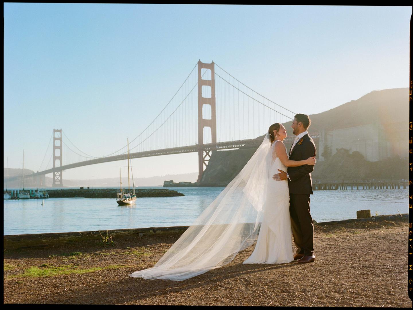 A couple embraces on a dirt path with the Golden Gate Bridge and a sailboat in the background.