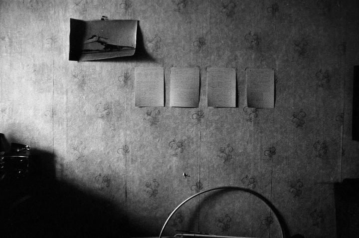 A black and white image shows a patterned wall with a small photograph and four pieces of paper taped to it.