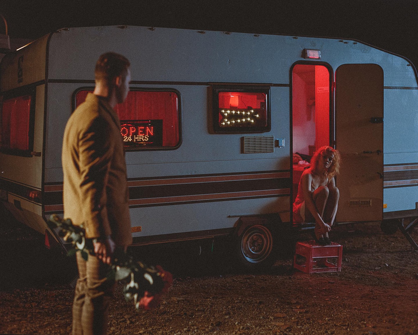 A man holding roses stands in front of a trailer with a woman sitting in its doorway, illuminated by red light at night.