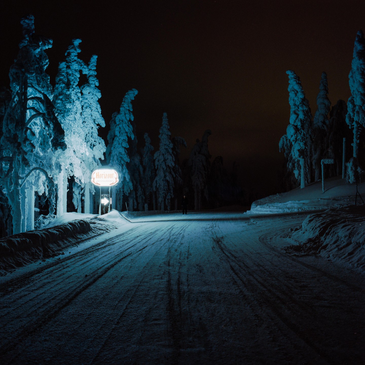 A snow-covered road at night leads to a glowing "Horizon" sign and a forest of snow-laden trees.