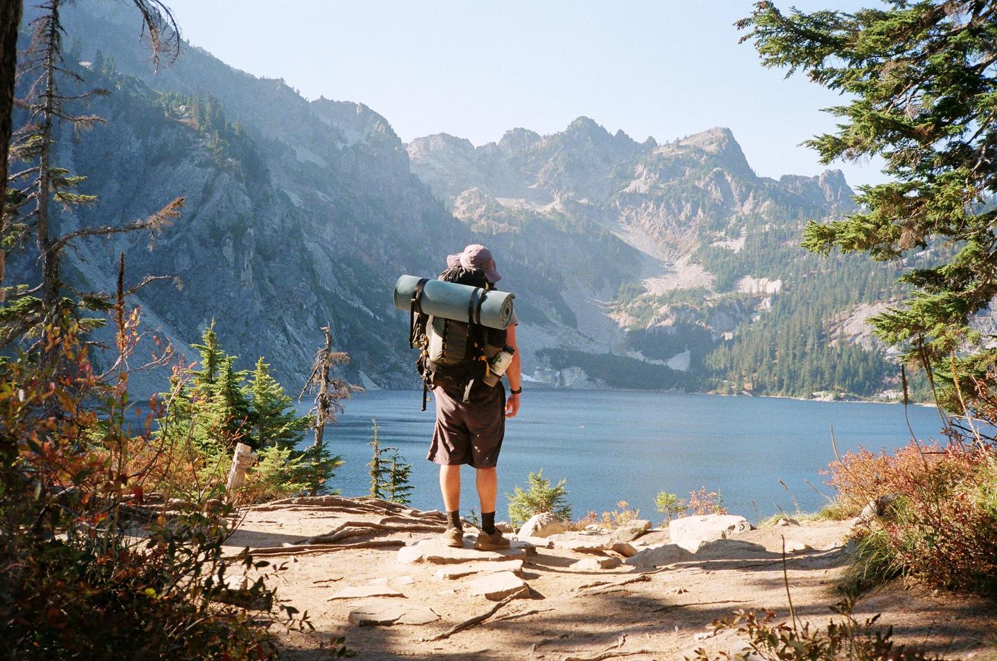 A hiker with a large backpack stands on a rocky path overlooking a lake surrounded by mountains and trees.