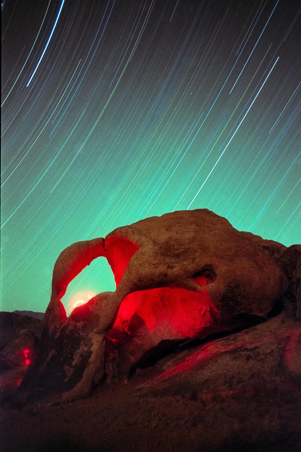 A rock formation with an arch is illuminated in red under a night sky with star trails.