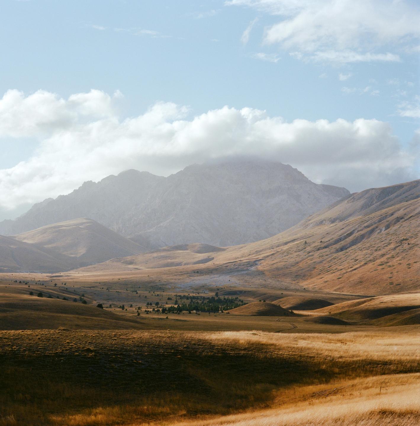 A vast landscape shows rolling hills of dry grass in the foreground and middle ground, with large mountains under a cloudy sky in the background.
