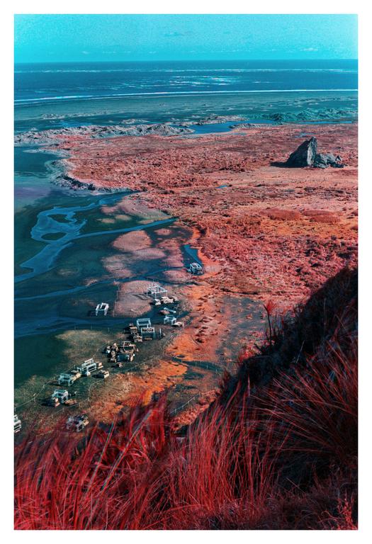 A high-angle landscape shows a body of water next to a red-hued land with a cluster of structures and a large rock formation.