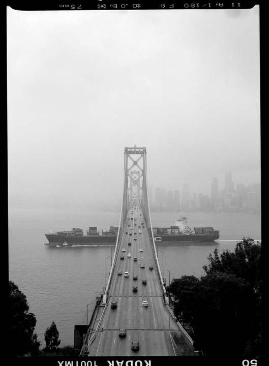 A cargo ship passes under a bridge with cars driving on it, with a city skyline in the background.