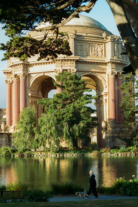 A woman walks a dog along a path next to a pond with a large domed building and trees in the background.
