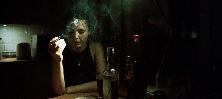 A woman sits at a table in a dark room, holding a lit cigarette with smoke rising around her head.